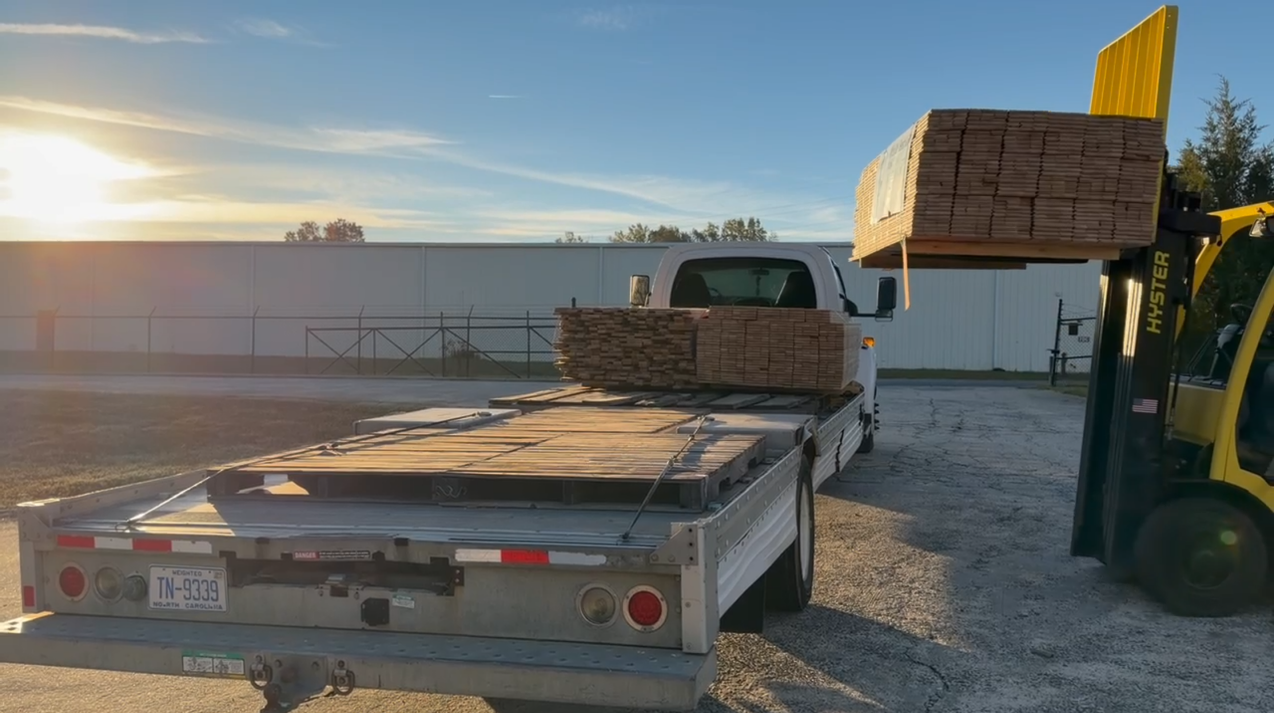Forklift holding a pallet of hardwood flooring beside a flatbed truck outside a warehouse.
